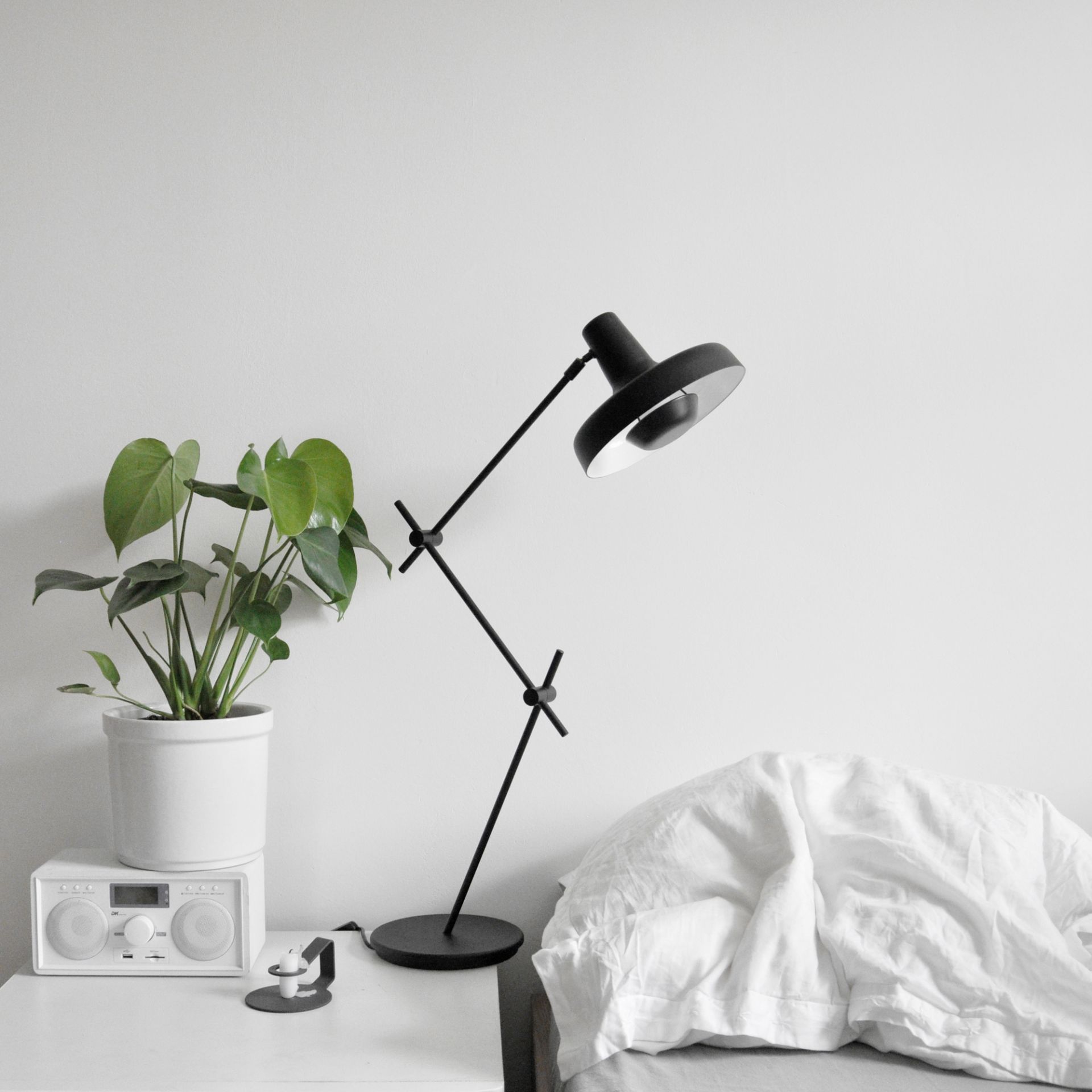 Nachttisch mit schwarzer Schreibtischlampe, Zimmerpflanze im weißen Topf und weißem Bettzeug, minimalistisches Design.