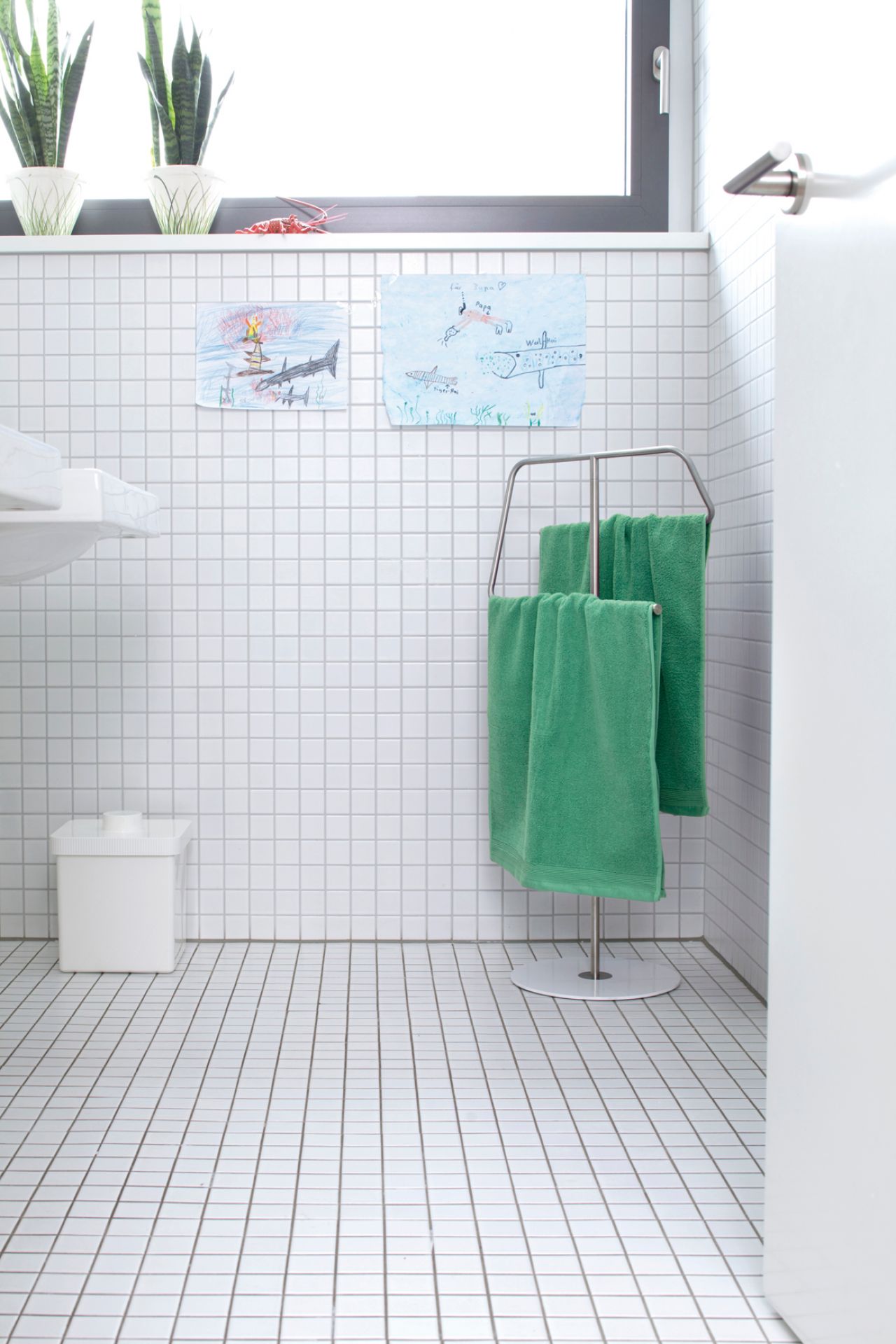 Badezimmer mit weißen Fliesen, Handtuchständer mit grünen Handtüchern und Kinderzeichnungen an der Wand.