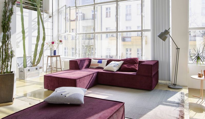 Burgunderrotes Ecksofa aus Cord mit Kissen in modernem Wohnzimmer mit großem Fenster und Pflanzen.