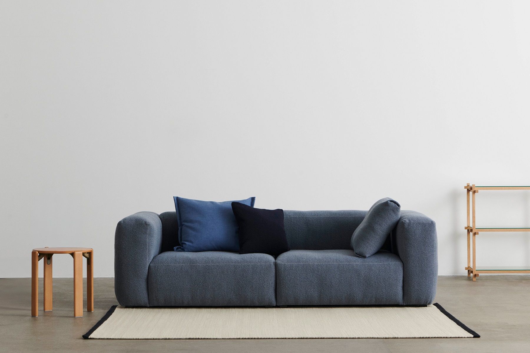 Blaues Sofa mit Kissen und ein Hay Woody Column Low Regal im Hintergrund vor einer weißen Wand.