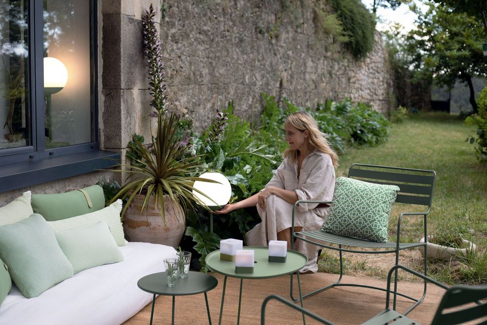 Frau entspannt im Garten auf Fermob Gartenmöbeln, darunter ein Surprising Sessel und ein Outdoor-Sofa mit Kissen.