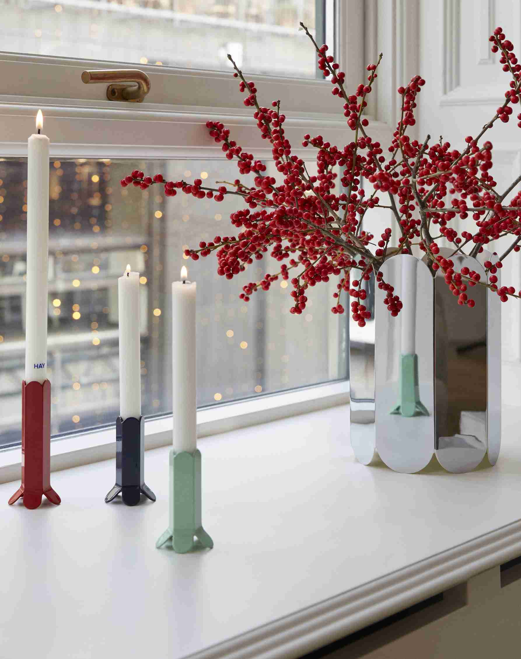 HAY Arcs Vase L Mirror: Silberne Design-Vase mit roten Beeren und Kerzenständern auf einer Fensterbank.