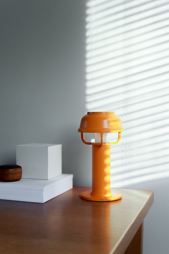 Orangefarbene Kori Tischleuchte von Artek auf einem Holztisch mit Büchern und Jalousien im Hintergrund.