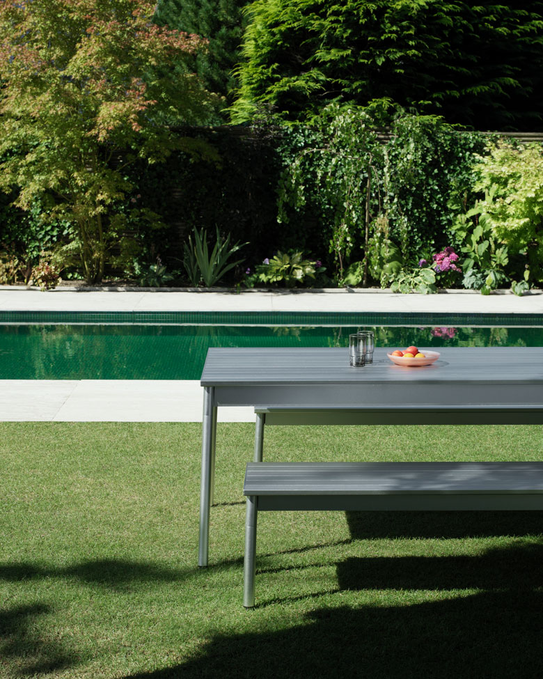 Normann Copenhagen Ex Tisch Outdoor mit Bank auf Rasen neben Pool, zwei Gläser und Obstschale auf Tisch.