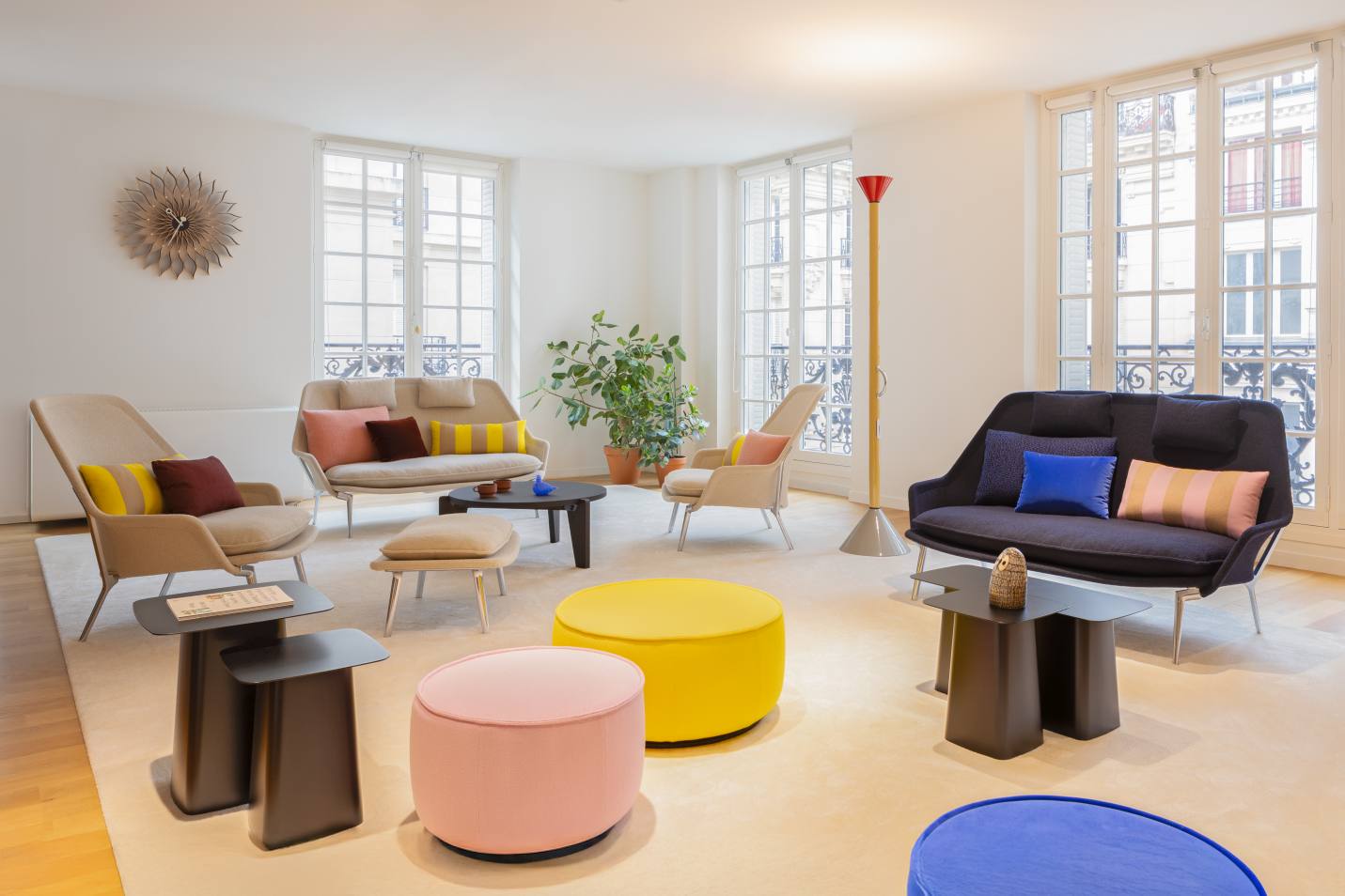 Helles Wohnzimmer mit Vitra Slow Sofa aus Stoff, Sesseln, Poufs und Beistelltischen auf einem Teppich.