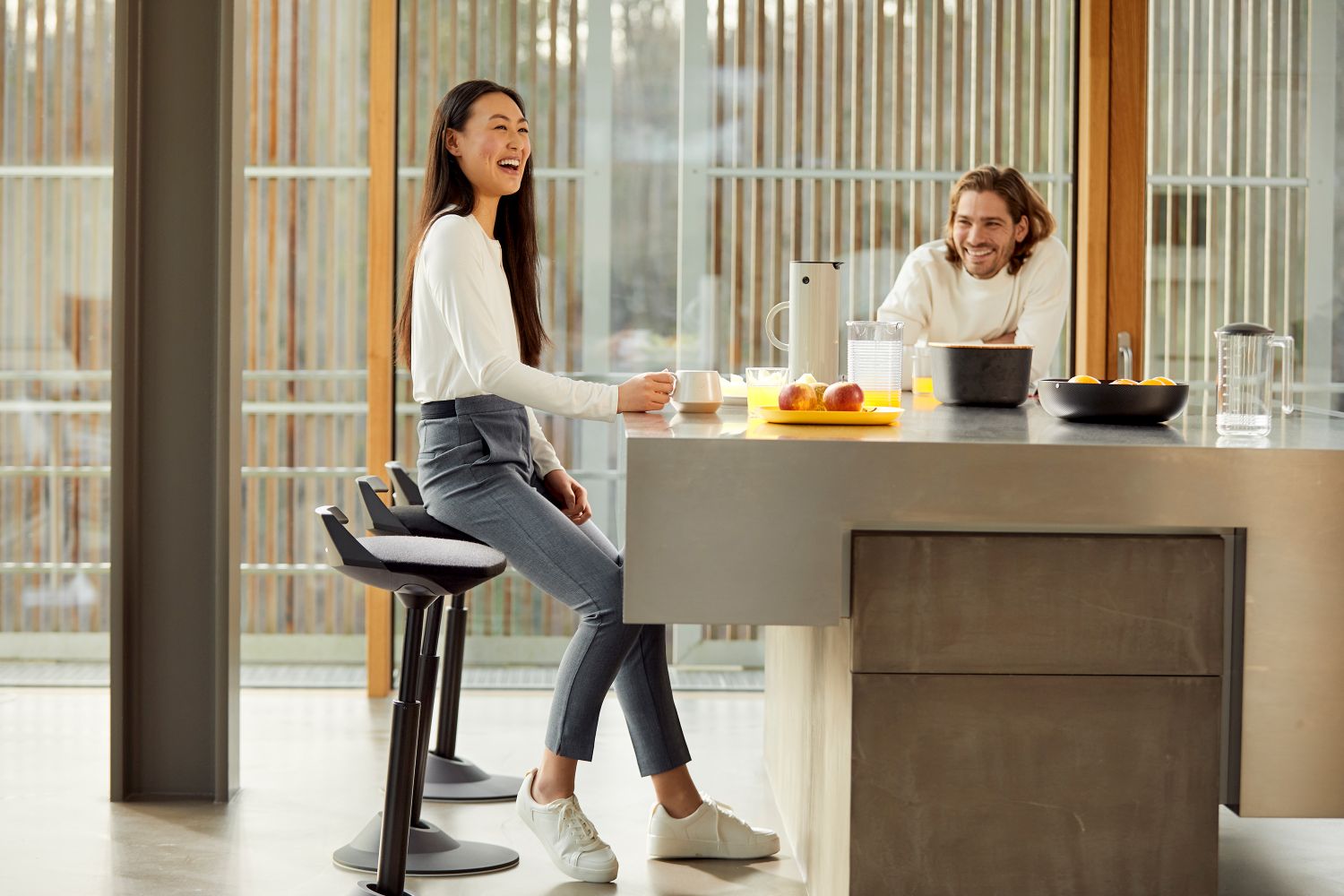 Frau sitzt lachend auf einem grauen Muvman Stehhocker am Küchentresen. Ergonomischer Sitzhocker für dynamisches Sitzen.