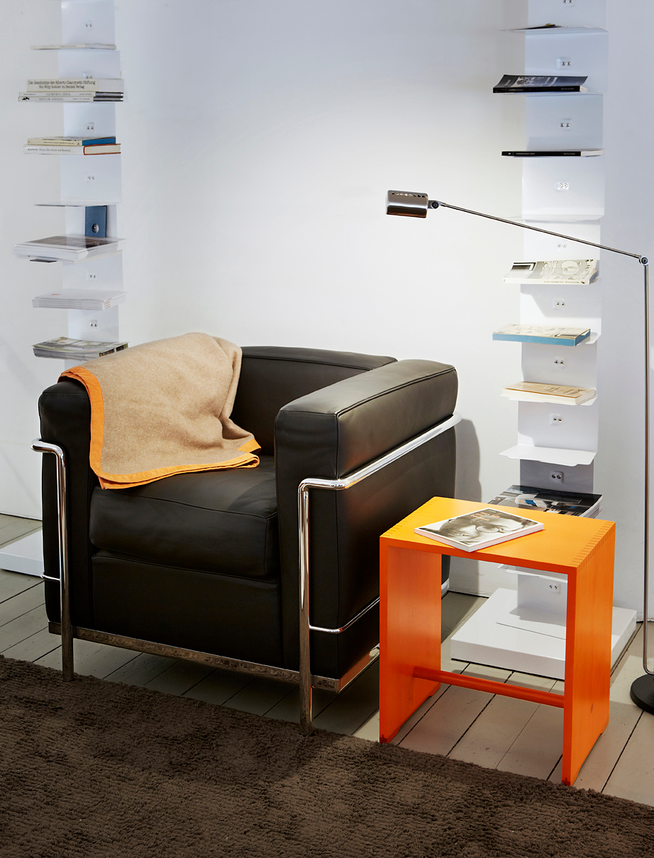 Leuchtoranger Ulmer Hocker als Beistelltisch neben schwarzem Sessel in modernem Wohnzimmer mit Bücherregalen und Stehlampe.
