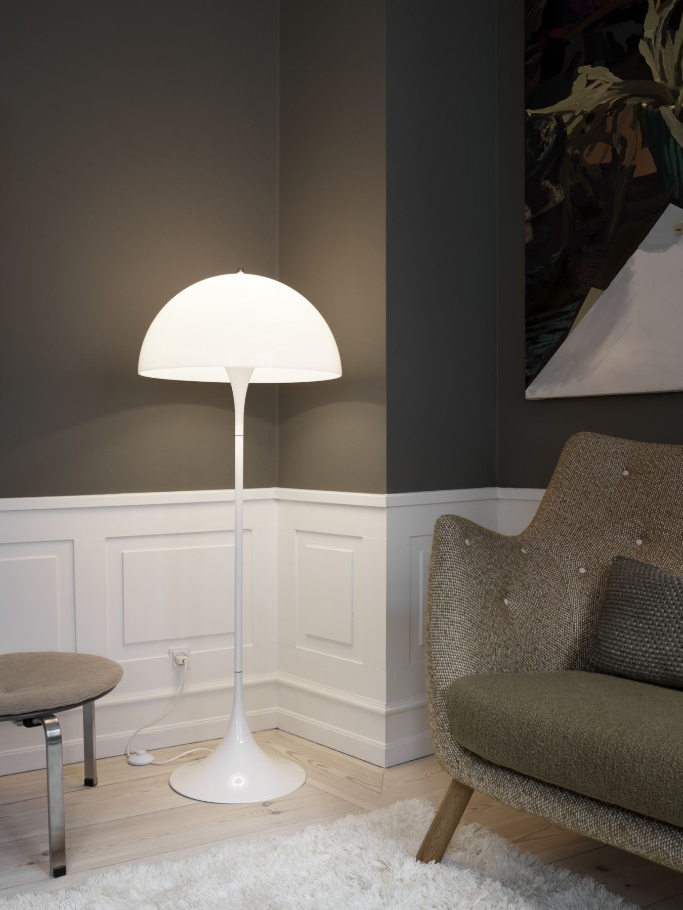 Moderne weiße Stehlampe im Interieur mit Sessel, Hocker und Kunstwerk an dunkler Wand.
