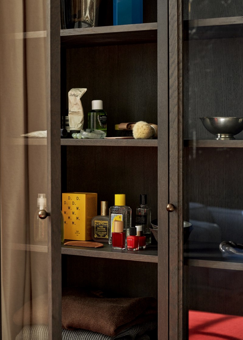AndTradition Trace SC88 Vitrine aus dunklem Holz mit Glasfront, gefüllt mit Kosmetikartikeln und Accessoires.