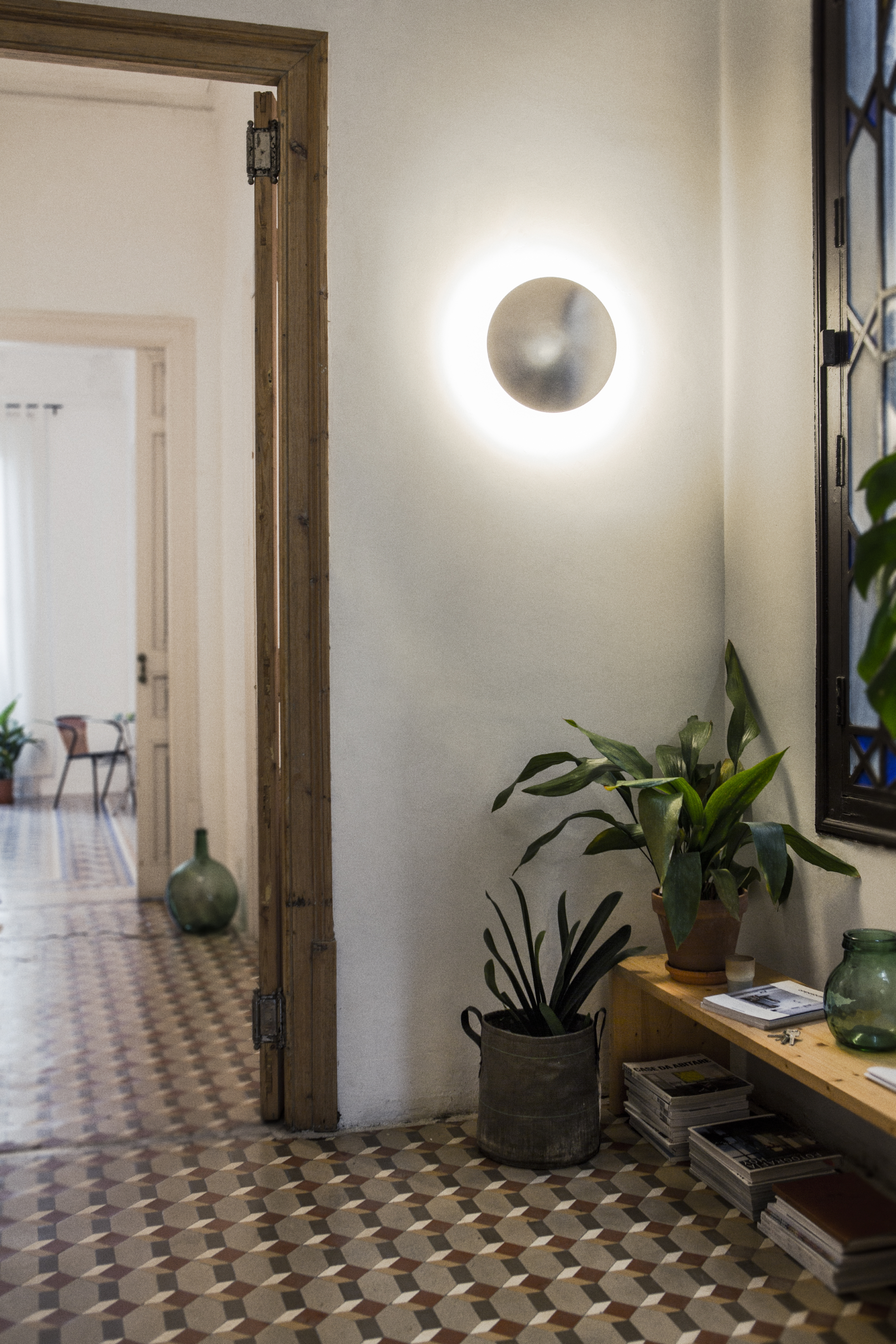 Helle Santa & Cole Disco Wandleuchte an weißer Wand im Flur mit Pflanzen.