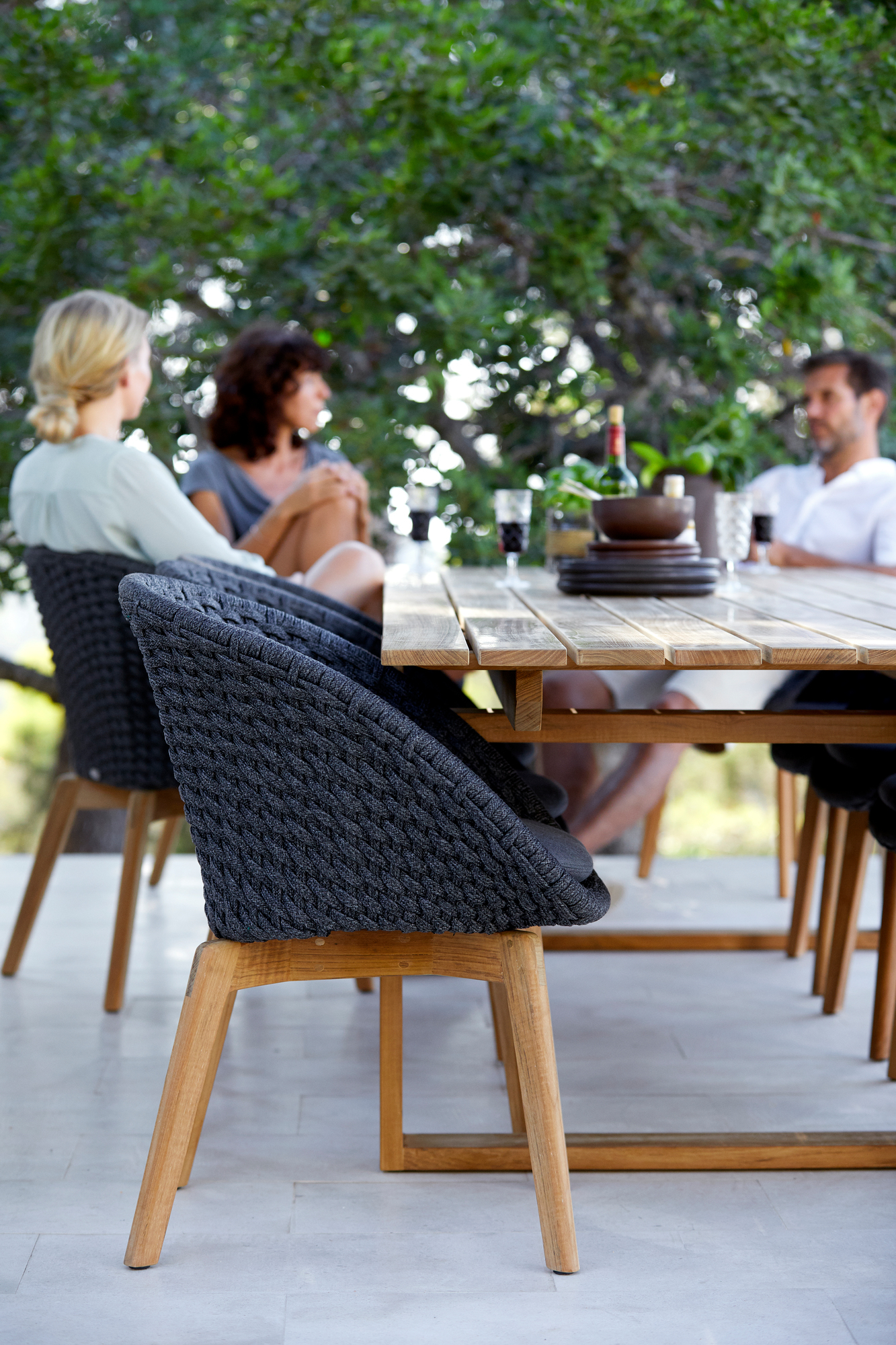Dunkelgrauer Cane-Line Peacock Outdoor Sessel mit Seilgeflecht und Holzbeinen am Esstisch.