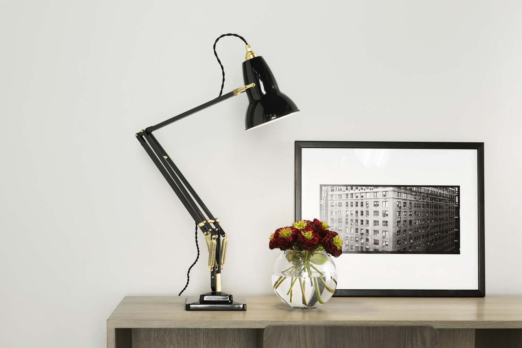 Schwarze Anglepoise Original 1227 Messing Tischleuchte auf einem Schreibtisch, neben einer Vase mit Blumen.