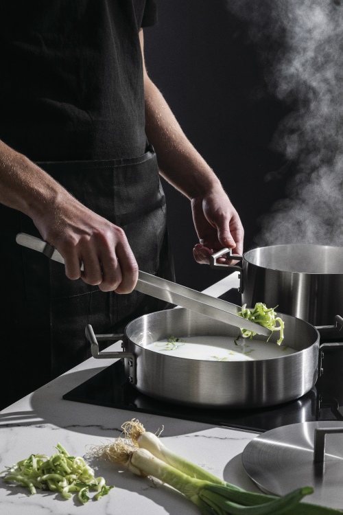 Alessi Convivio Kochtopfset: Person kocht mit Zange Gemüse in einem Topf auf einem Induktionsherd.