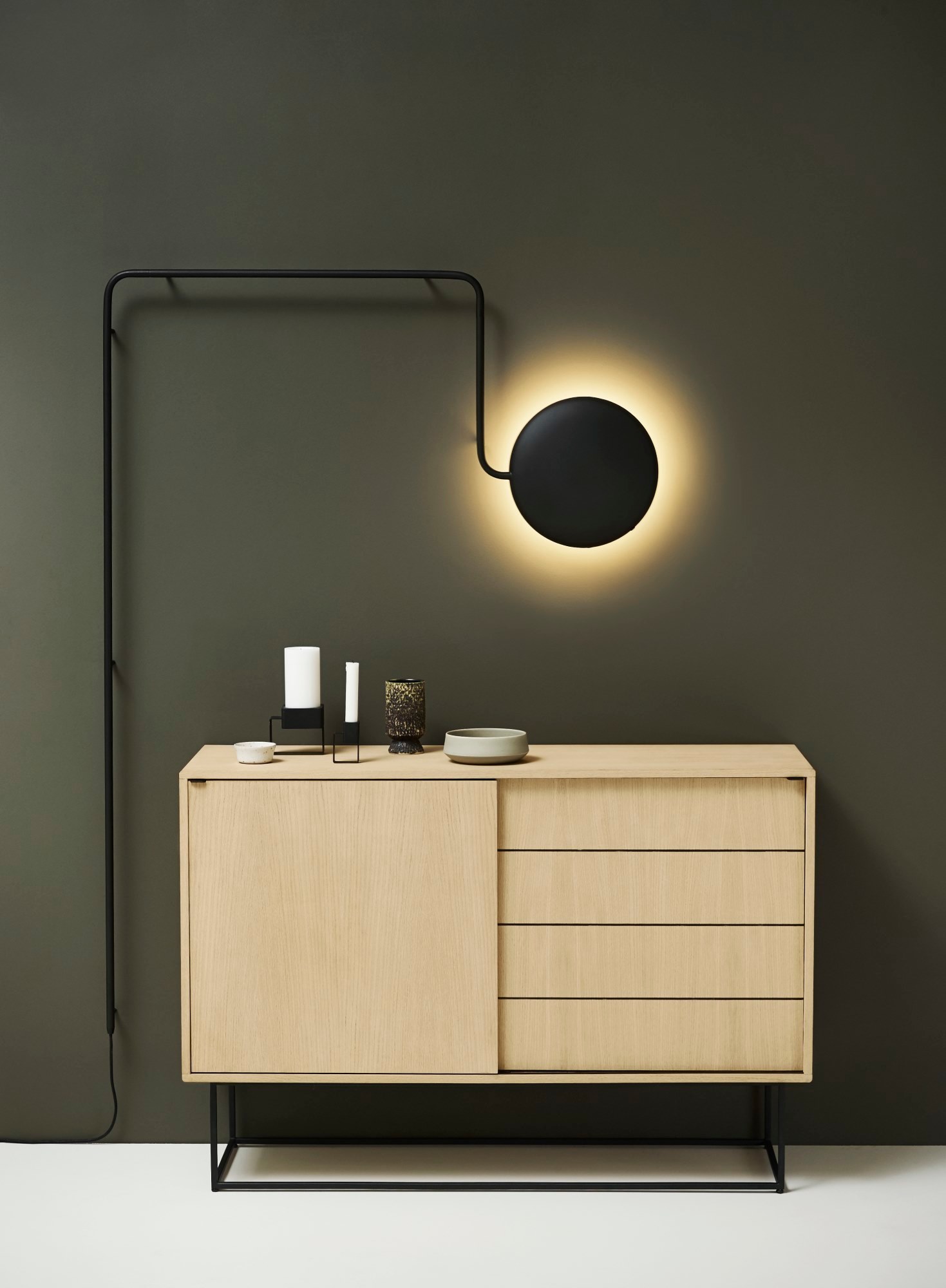 Schwarze Mercury Wandleuchte von Woud an grüner Wand über Kommode mit Deko-Elementen.