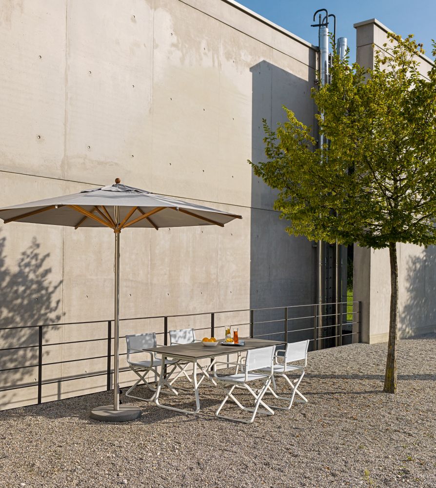 Ahoi Tisch Outdoor von Weishäupl mit Sonnenschirm und vier Stühlen auf Kiesfläche vor Betonwand.
