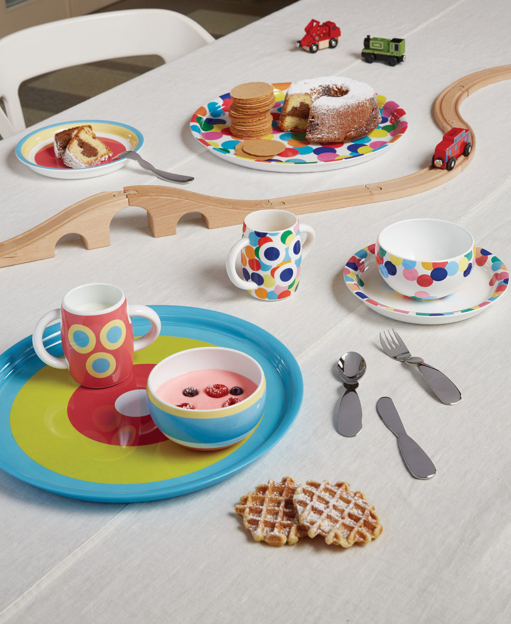 Alessini Kindergeschirr-Set: Bunte Teller, Tassen und Edelstahl-Besteck für Kinder auf einem Tisch.