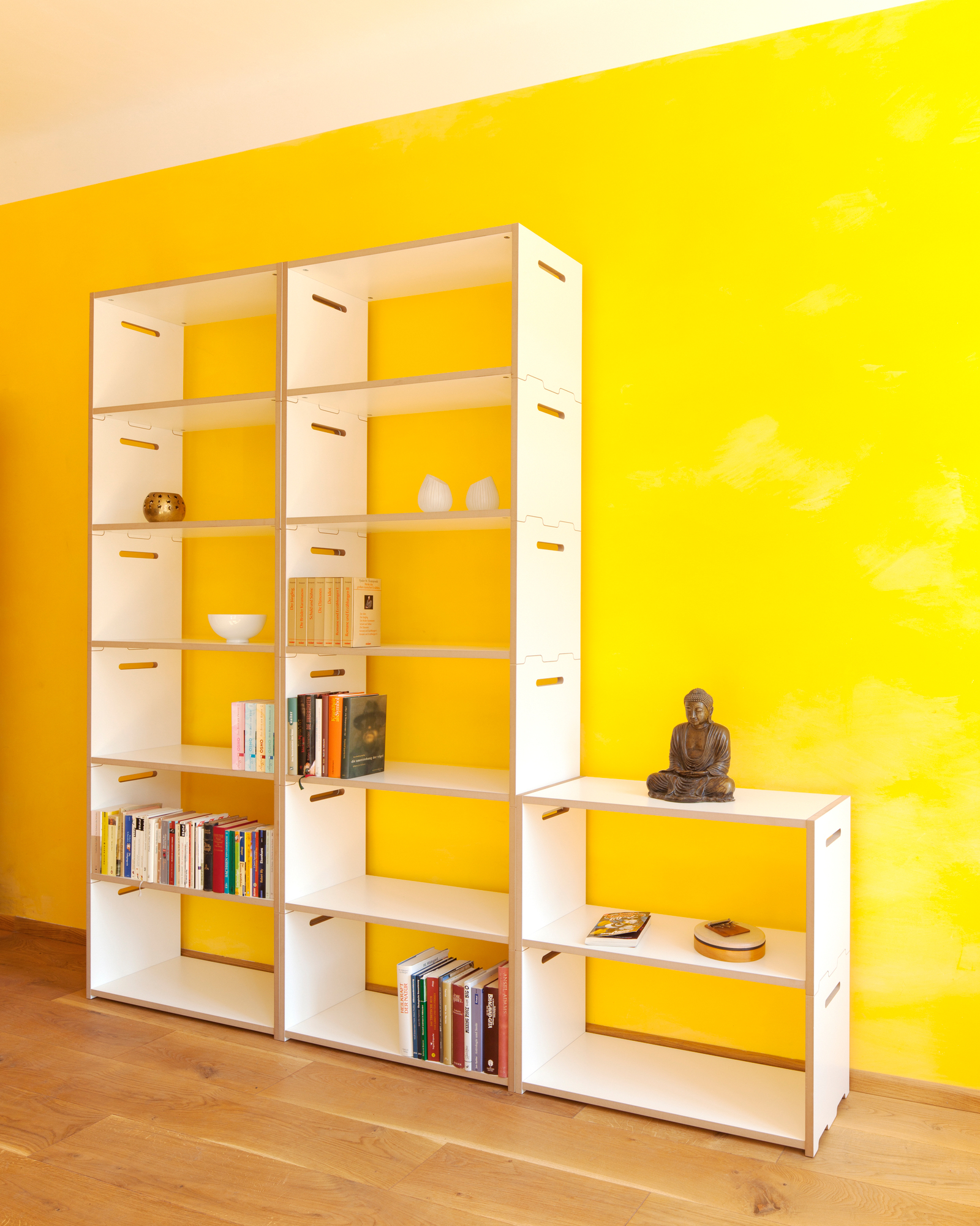Tojo Hochstapler Regal mit Anbaumodul, weißes modulares Regalsystem vor gelber Wand, Bücher und Deko.