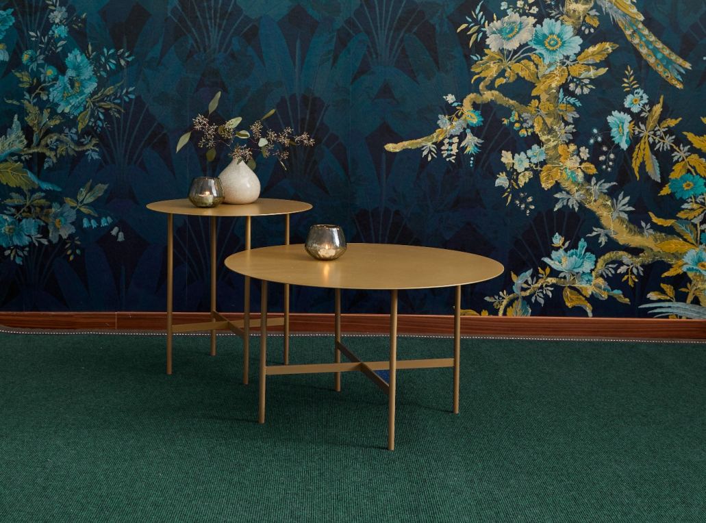 Zwei goldene Couchtische mit Kerzenhaltern vor einer floralen Tapete auf grünem Teppich.