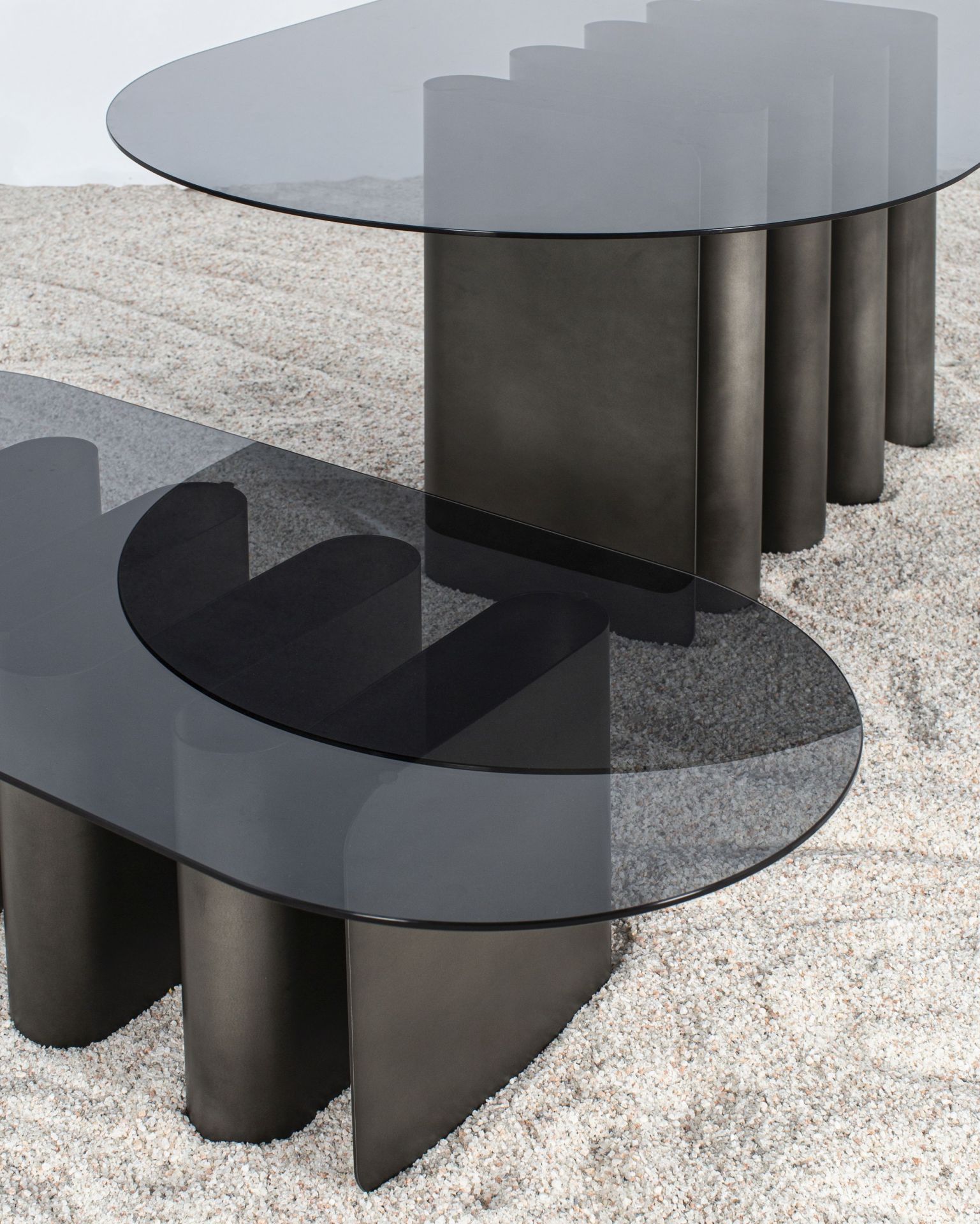Zwei moderne Esstische mit grauen Glasplatten und Metallgestell auf hellem Kiesboden.