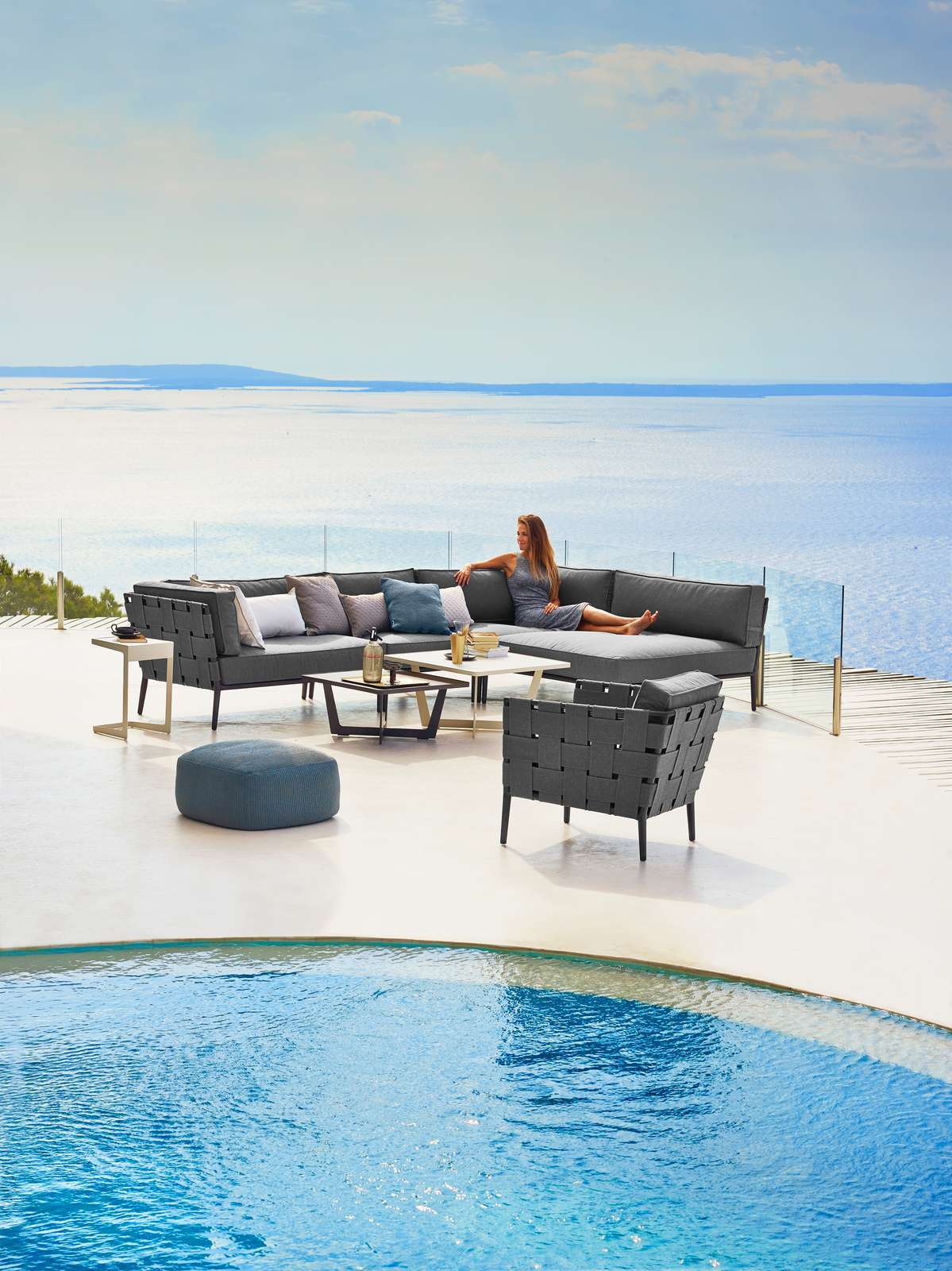 Frau entspannt auf Cane-Line Conic Outdoor Lounge Sofa mit Meerblick, graue Gartenmöbel mit Kissen.
