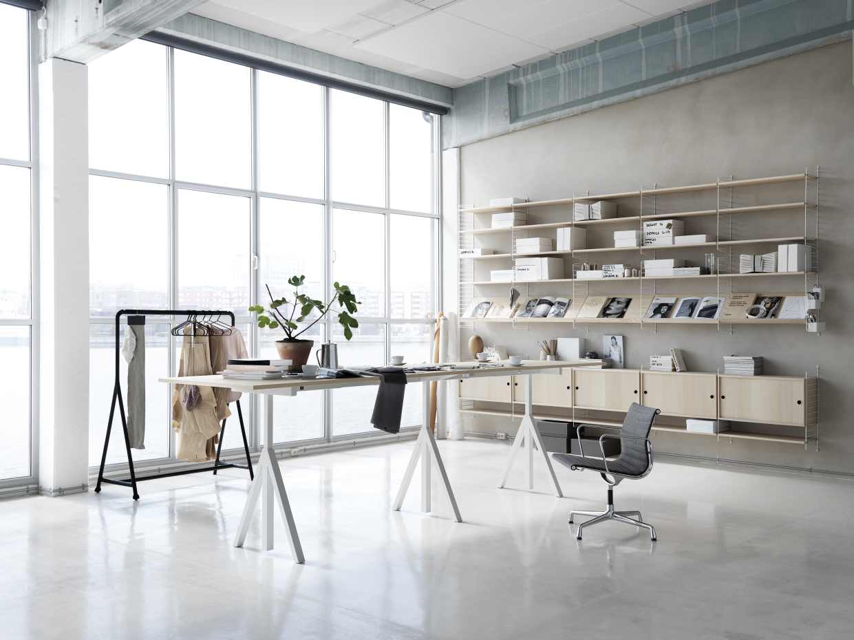 Heller Arbeitsbereich mit Wordesk Table Schreibtisch von String Furniture, Regalsystem, Kleiderständer und großem Fenster.
