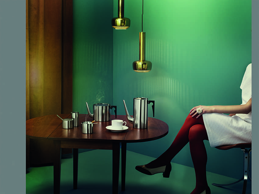 Edelstahl Tee- und Kaffeeset auf Holztisch, goldene Hängelampen im Hintergrund, Frau im weißen Kleid.