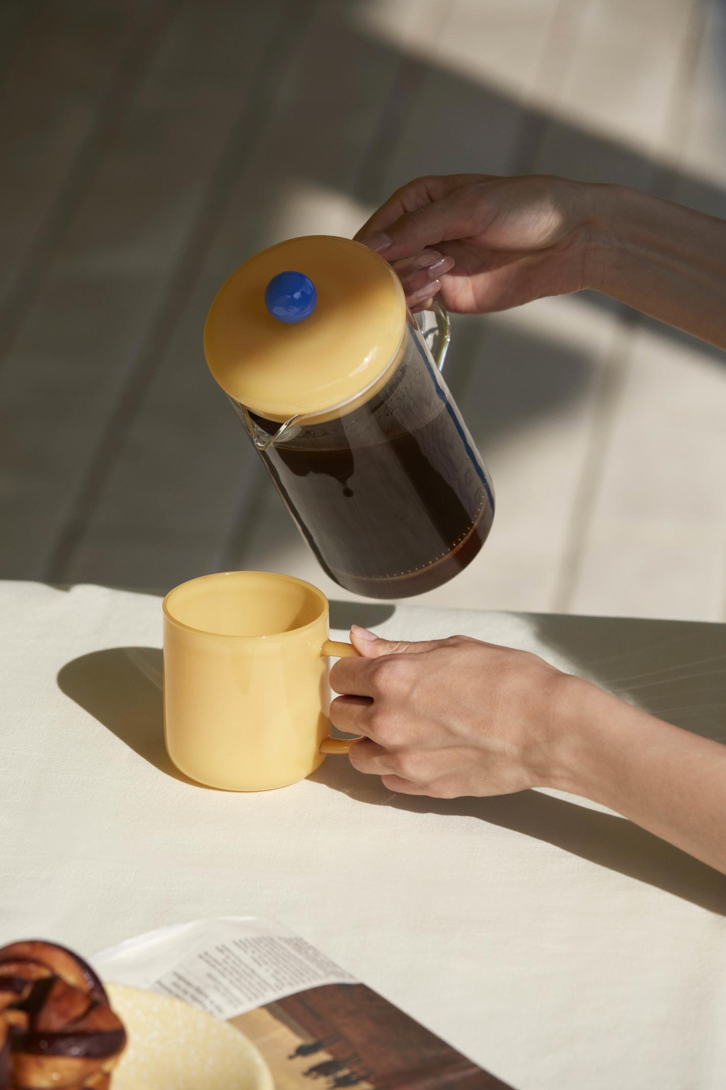 Hände gießen Kaffee aus einer gelben French Press in eine passende Tasse. HAY Kaffeebereiter.