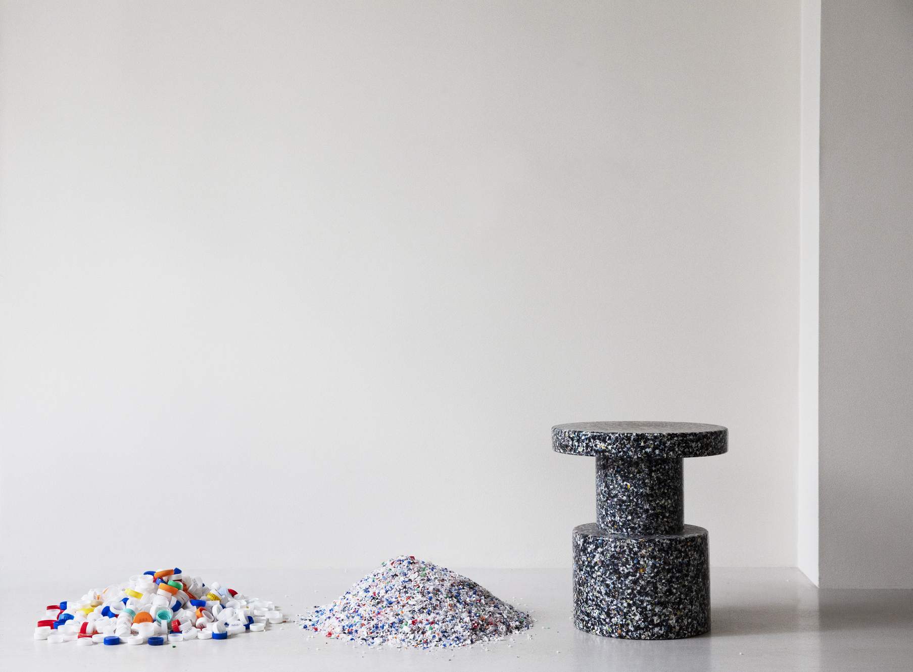 Normann Copenhagen Bit Stool Hocker aus recyceltem Kunststoff, neben Rohmaterialien wie Kunststoffgranulat und Deckeln.