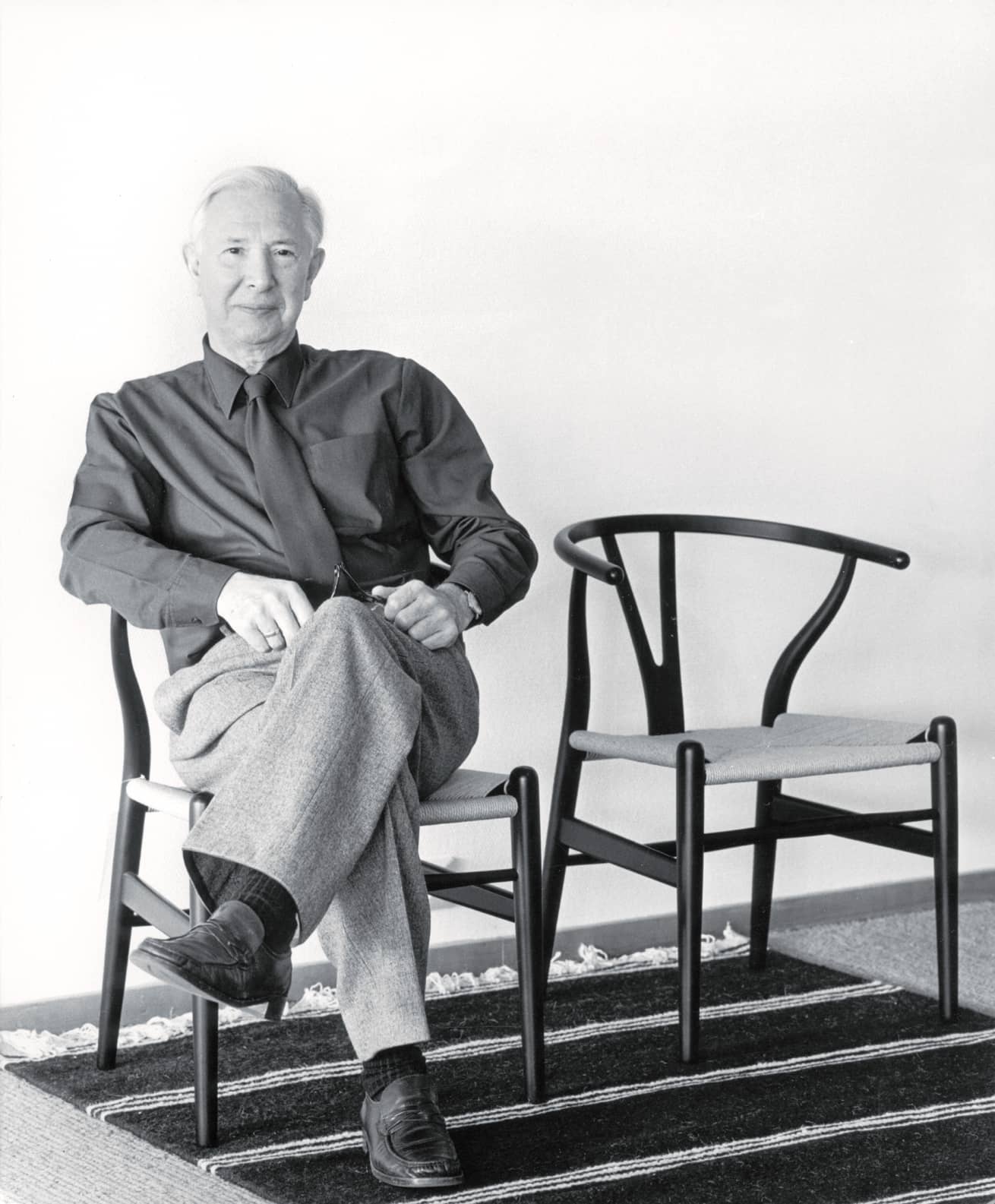 Schwarz-Weiß-Foto: Mann sitzt auf einem Wishbone Chair von Hans Wegner, davor ein weiterer Stuhl.