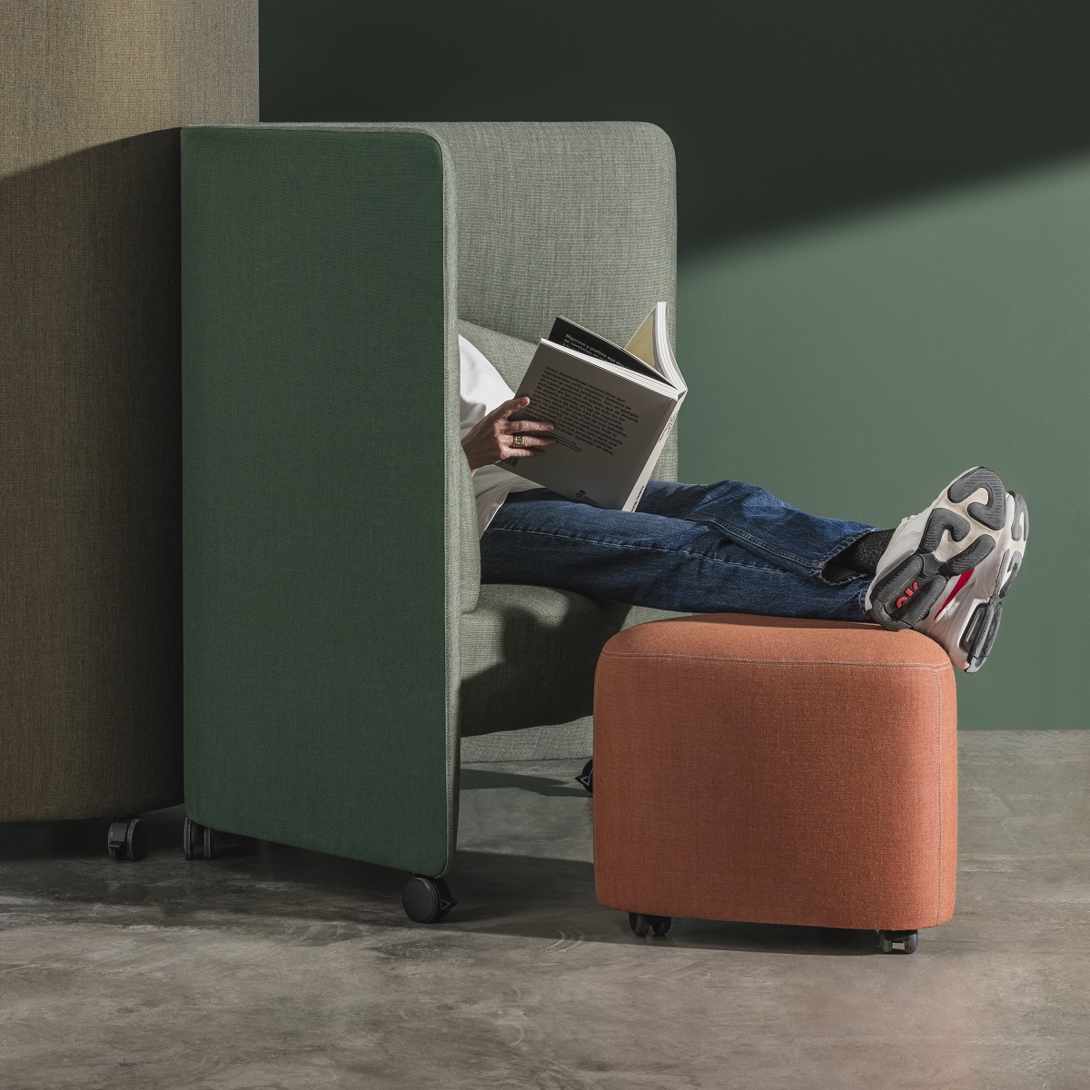 In Out Office Loungesessel mit grünen Akustikpaneelen und orangefarbenem Hocker, Person liest entspannt.