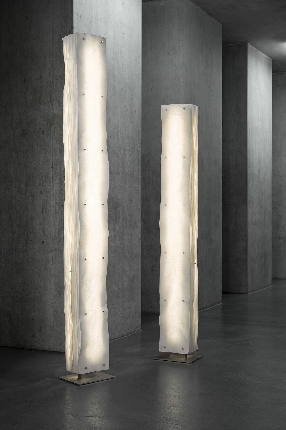 Zwei leuchtende Totem Stehleuchten im Stele-Design vor einer Betonwand, moderne Innenraumbeleuchtung.