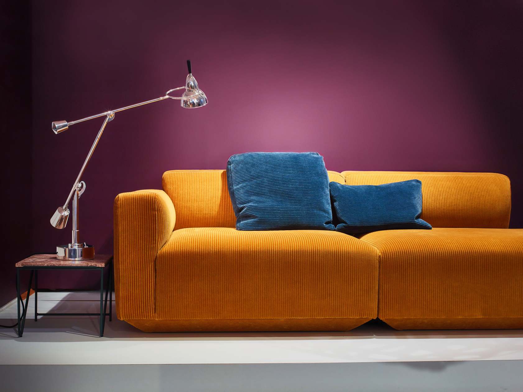 Goldenes Cord Sofa mit blauen Kissen und silberner Lampe vor einer lila Wand.