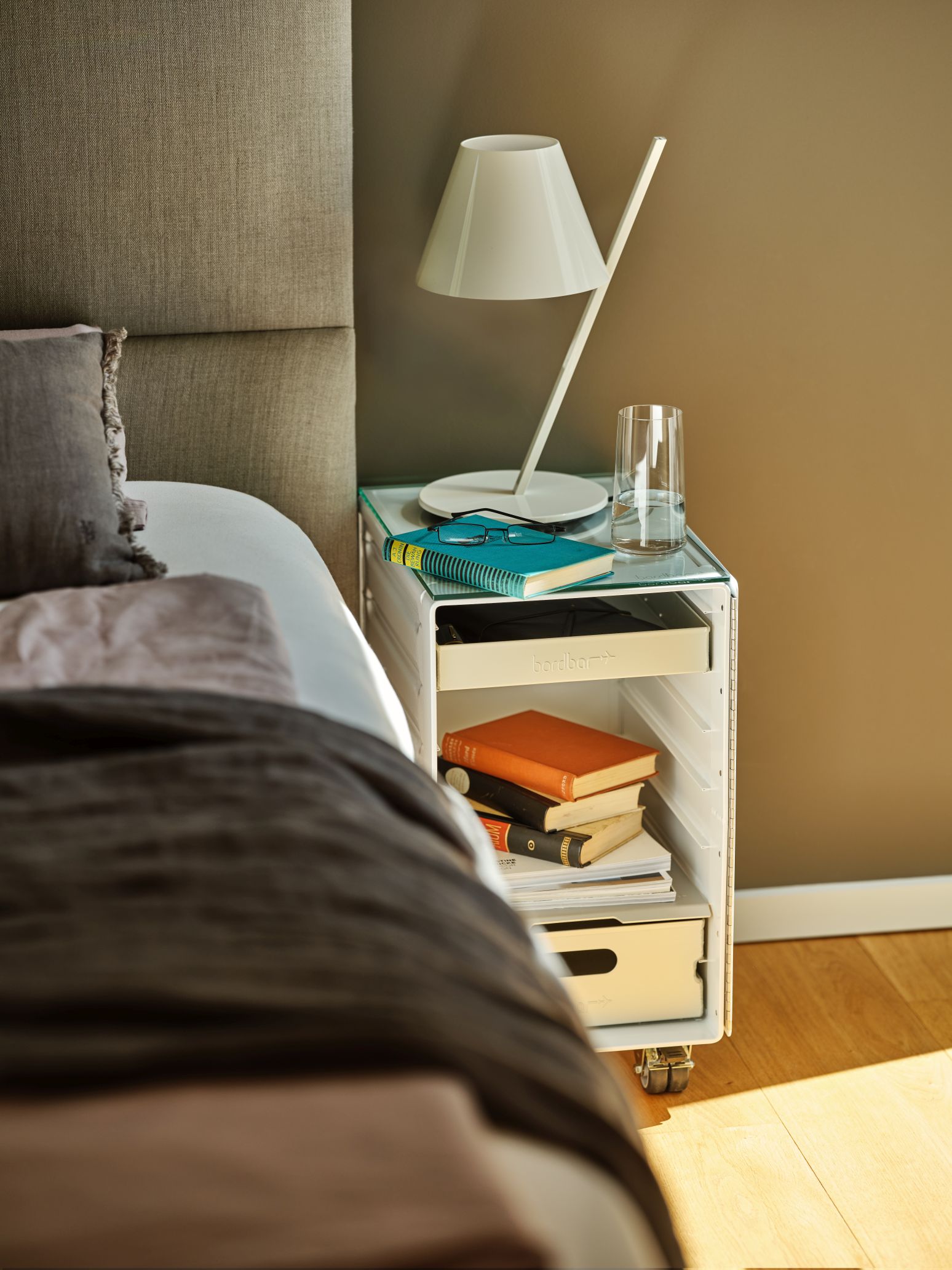 Box Nachttisch mit Lampe, Brille, Notizbuch und Bücher als Zubehör neben einem Bett.