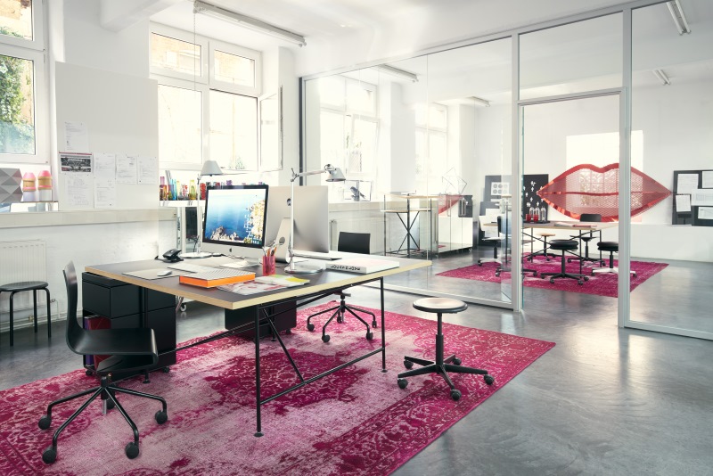 Helles modernes Büro mit Schreibtischen, Computern, schwarzen Bürostühlen und pinkfarbenen Teppichen für eine kreative Arbeitsumgebung.
