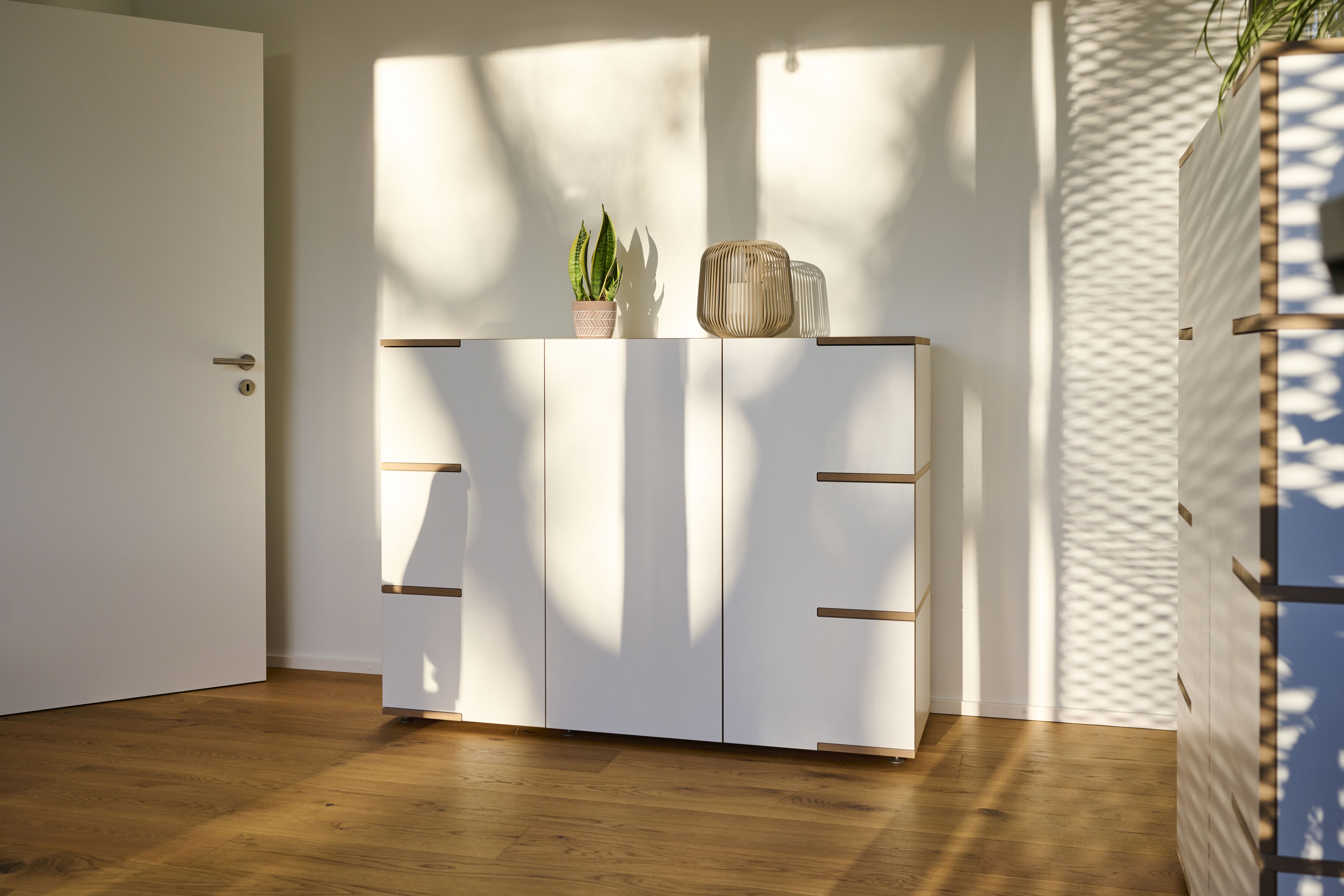 Weißes Stau Sideboard von Tojo mit Holzdetails, dekoriert mit Pflanze und Windlicht im hellen Raum.