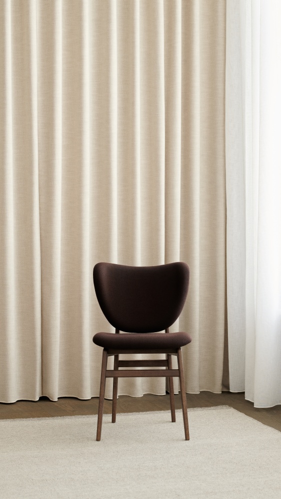 Vollgepolsterter, brauner Elephant Dining Chair Stuhl von NORR11 vor beigen Vorhängen, geeignet für Esszimmer und Wohnbereich.