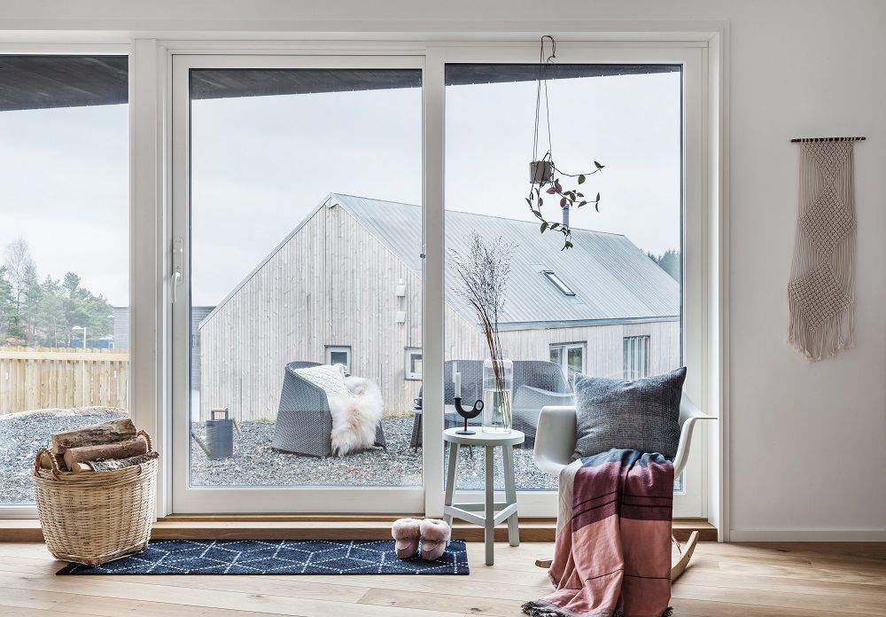 Helle Raumansicht mit schwarzer Heymat Fußmatte, Holzbündel im Korb und Blick durch die Fensterfront.
