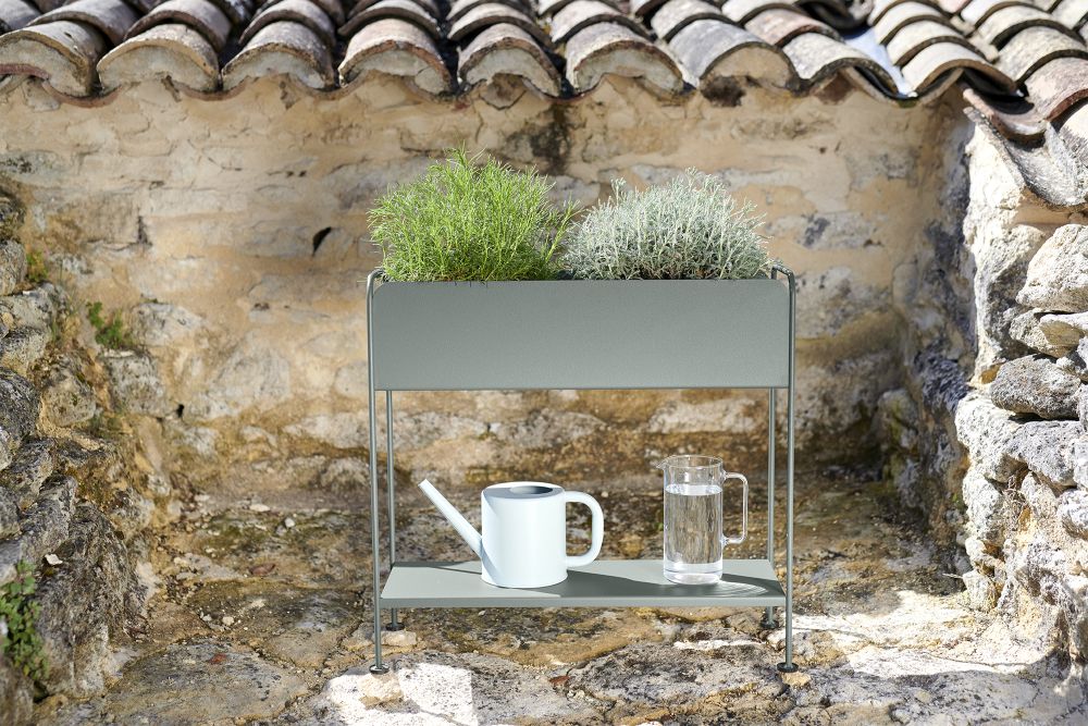 Picolino Übertopf Outdoor von Fermob mit Pflanzen, Gießkanne und Wasserkanne vor Steinwand.