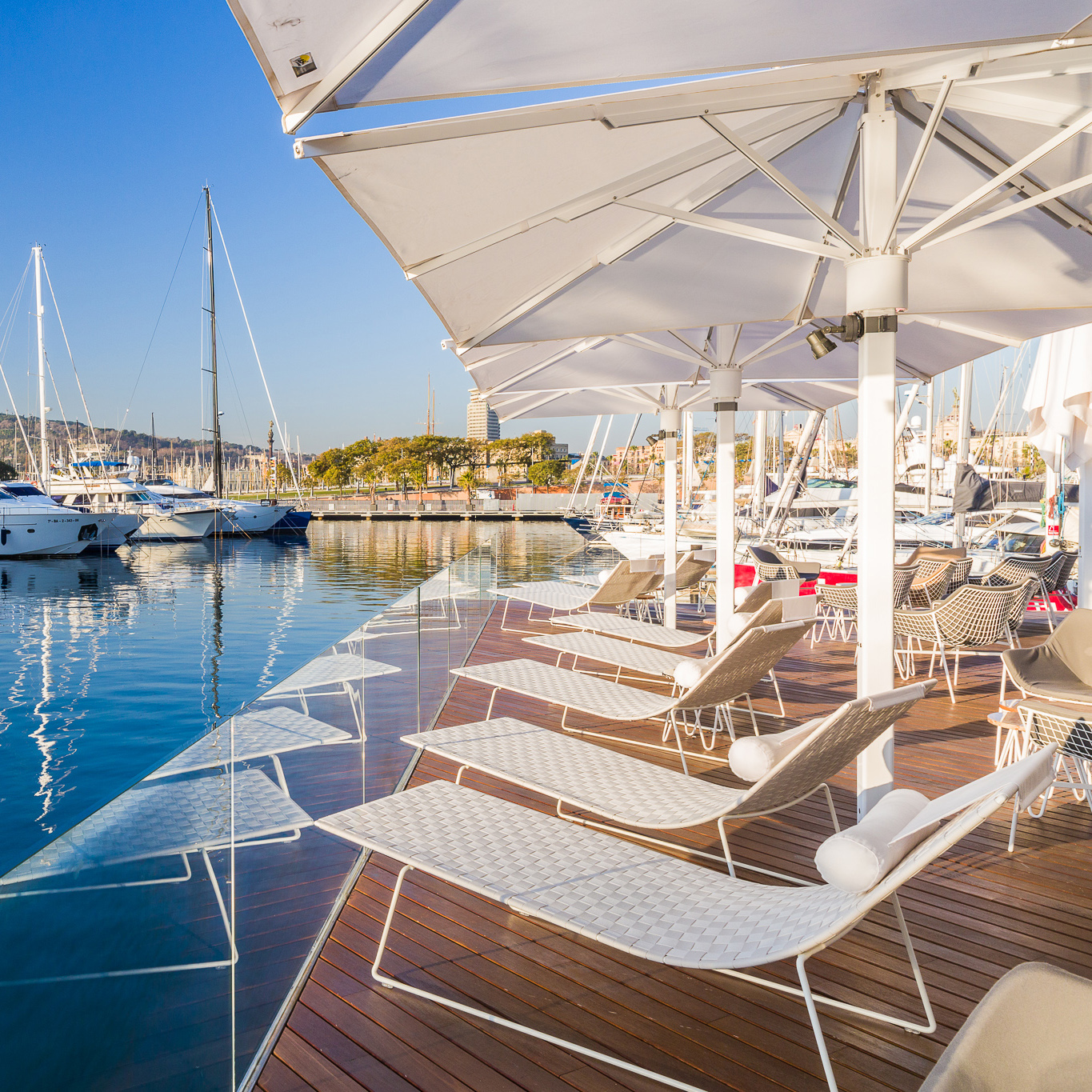 Weiße Trenza Chaise Lounges von Andreu World auf Holzdeck mit Blick auf Yachthafen und Sonnenschirme.