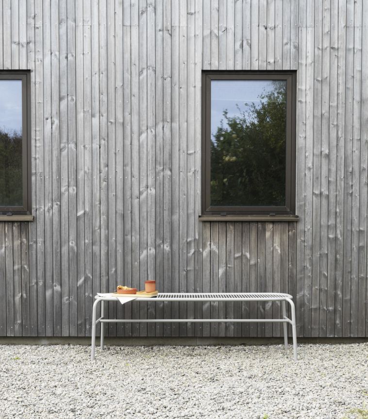 Normann Copenhagen Vig Bank, eine graue Outdoor-Bank vor einer Holzwand mit zwei Fenstern.