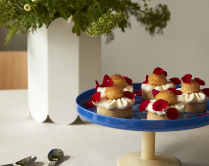 Fünf Mini-Baba-Kuchen auf blauem Tortenständer mit Rosenblättern, neben weißer Vase mit Blumen.
