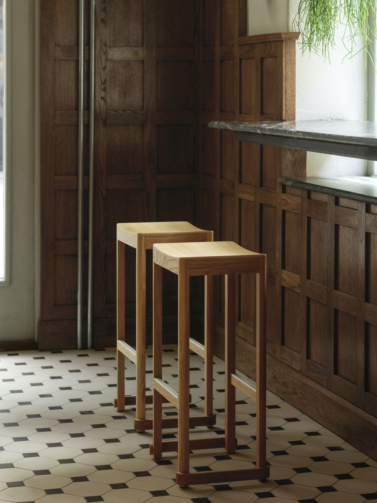 Zwei Atelier Bar Stool Barhocker aus hellem Holz vor dunkler Holzvertäfelung und Fliesenboden.