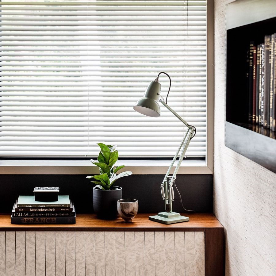 Grüne Schreibtischlampe im Retro-Design auf einem Holzregal mit Büchern, Pflanze und Jalousien im Hintergrund.
