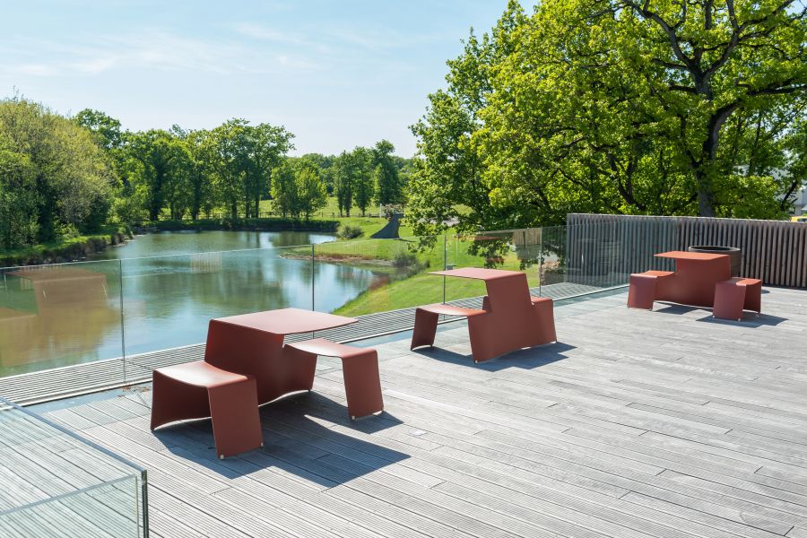 Roter Outdoor-Picknicktisch mit Bänken aus Metall auf Holzterrasse mit Blick auf See und grüne Landschaft.
