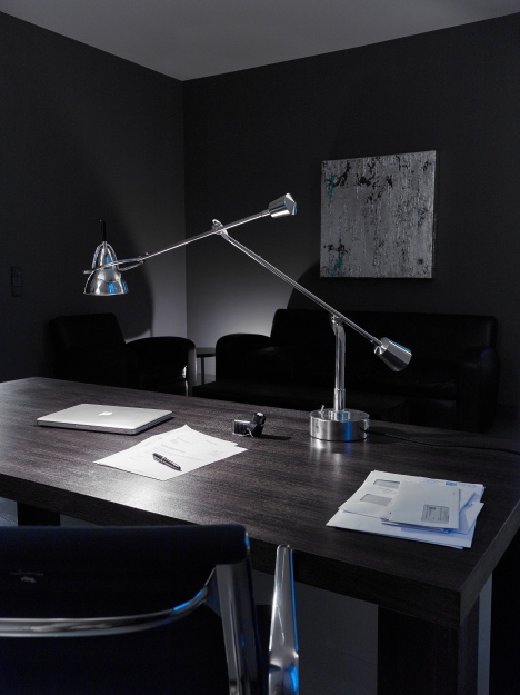 Moderne Schreibtischleuchte aus Chrom auf dunklem Holztisch mit Laptop, Papier und Stift im Büro.