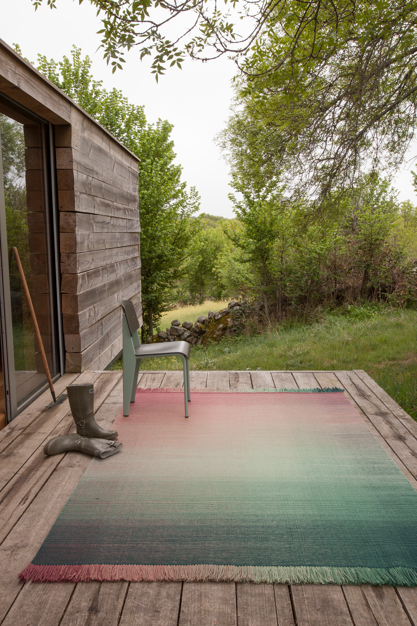 Nanimarquina Shade Teppich im Freien auf Holzterrasse mit Stiefeln und Stuhl, Farbverlauf von Rosa zu Grün.