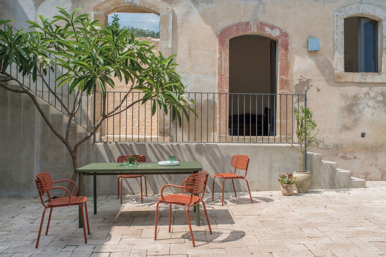 Gartenmöbel Set: Grüner Tisch und orangefarbene Stühle im mediterranen Außenbereich mit Baum.