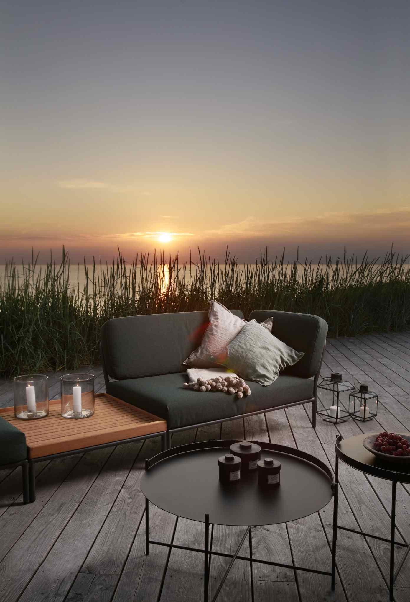 Houe Level Outdoor Couchtisch aus Teakholz neben Sofa auf Holzterrasse mit Meerblick bei Sonnenuntergang.