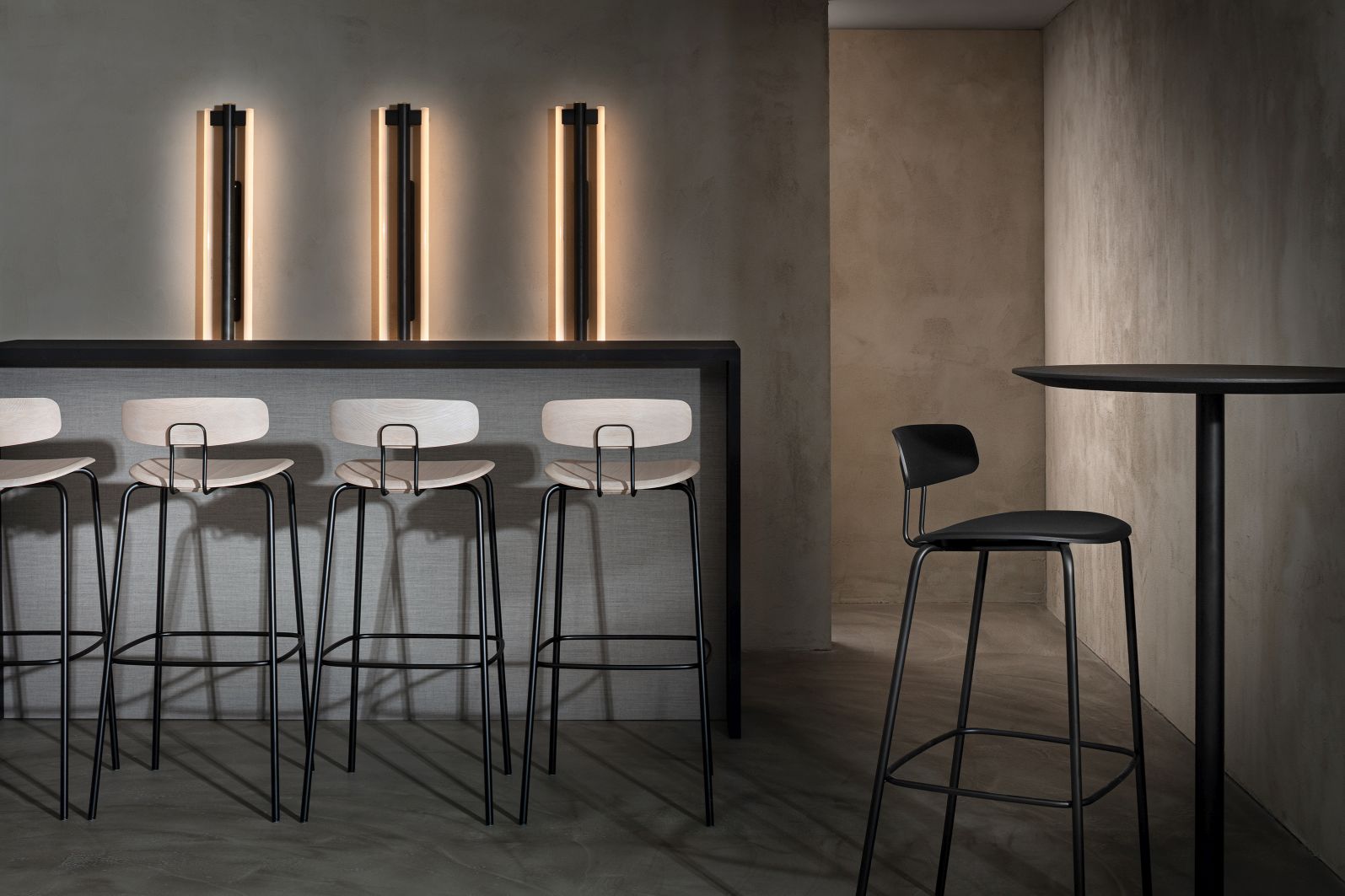 Okito Barstool Barhocker von Zeitraum, helle Holzsitze mit schwarzem Metallgestell vor grauer Wand.