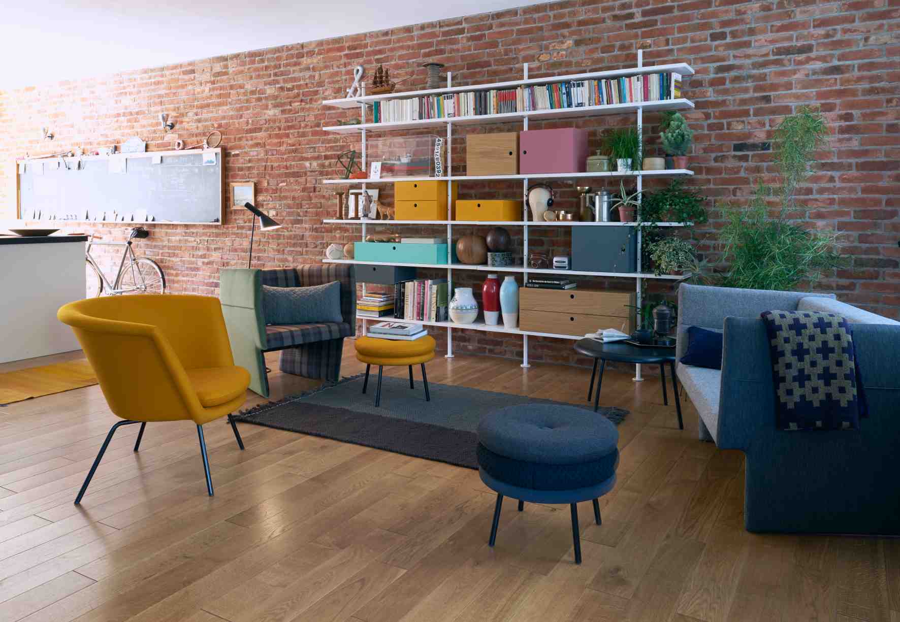 Wohnzimmer mit weißem Eiermann Regal mit Eichenholz-Elementen, Bücher und Deko vor einer Backsteinwand.